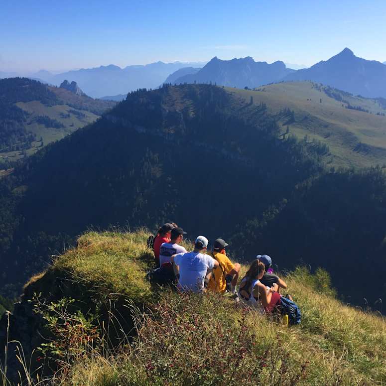 A group of student sitting on a grassy hill overlooking a view of mountains.
